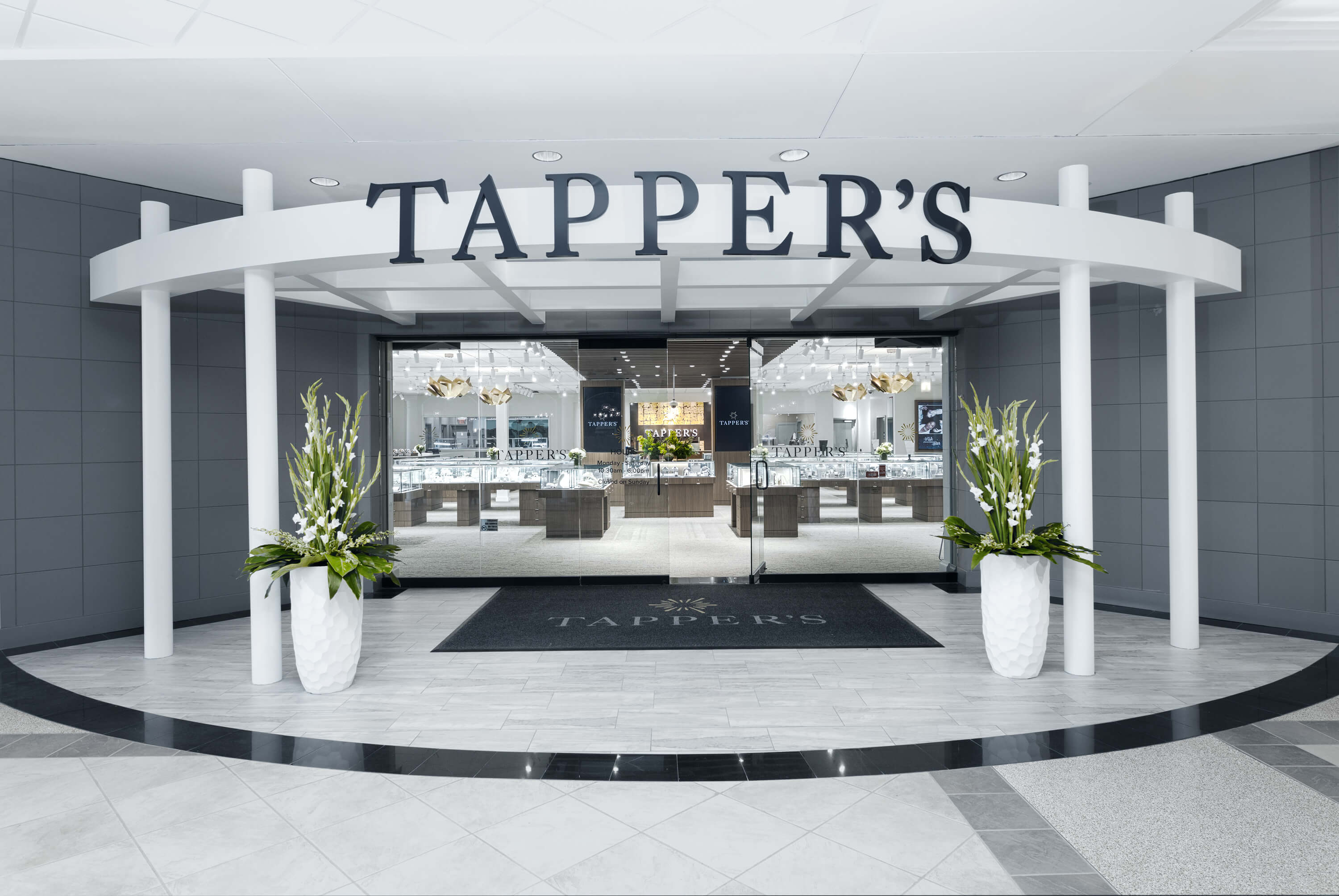 Tapper's West Bloomfield Entrance in Orchard Lake Mall. Gray tiled wall with white pillars frame the Tapper's logo. Tall white planters with fresh green floral accent the space.