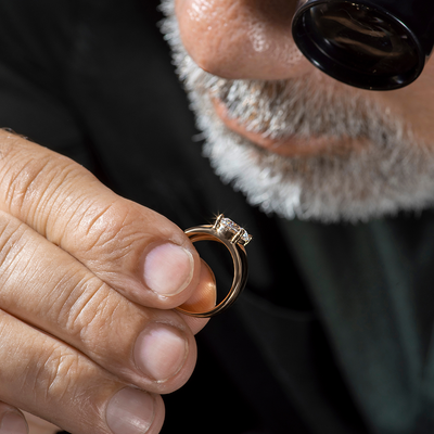 Man with white and grey beard examining a diamond gold ring with a jeweler's loop.