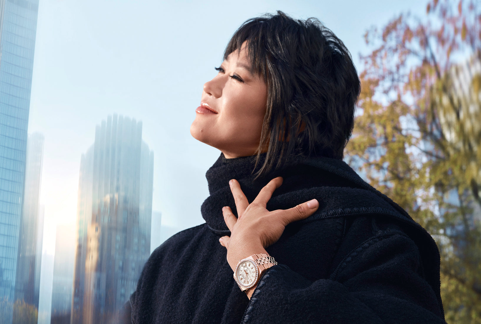 Person in a black coat wearing a rose gold Rolex watch on their wrist, set against a cityscape and autumn trees.