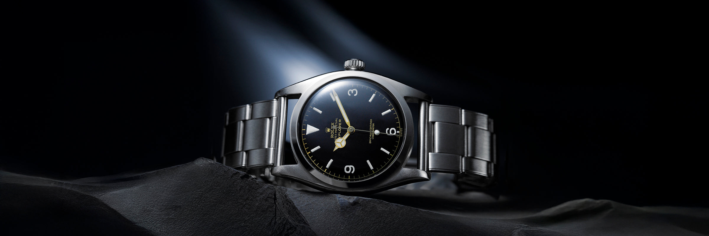 Vintage Rolex Explorer watch with black dial, luminous markers, and stainless steel bracelet shown under a spotlight on dark rocks.