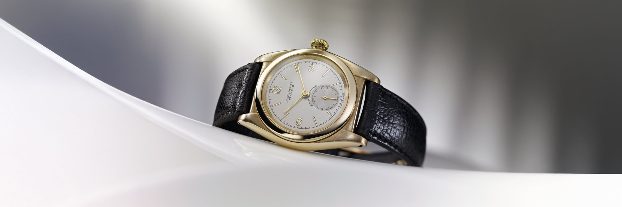 Vintage gold Rolex Oyster Perpetual wristwatch with white dial and black leather strap on a light background.