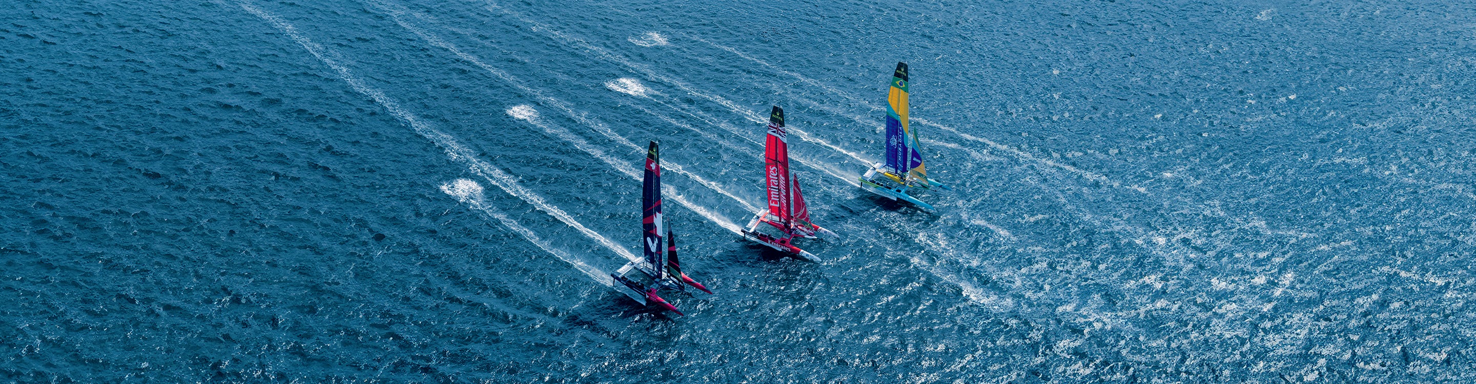 Aerial view of three high-speed sailboats racing on blue ocean waters during the Rolex SailGP Championship.